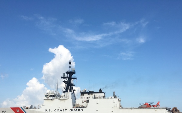 A U.S. Coast Guard National Security Cutter bearing hull number 754 is anchored or underway on calm, open blue water under a partly cloudy sky. The large white cutter displays the distinctive Coast Guard diagonal red-and-blue racing stripe on its bow. A red helicopter is visible on the aft flight deck, indicating the vessel's aviation capability.