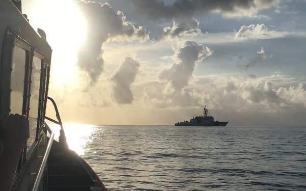 The image shows a maritime scene taken from the deck of a small vessel, looking out across calm open ocean toward a larger ship on the horizon. The scene is captured during either sunrise or sunset, with the sun low on the horizon creating a golden glare on the water. A larger vessel, consistent in silhouette with a Coast Guard or naval cutter, is visible in the middle distance.