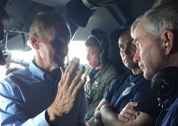 A group of individuals are gathered inside what appears to be a military or Coast Guard aircraft cockpit/cabin area. A civilian in a blue button-down shirt is gesturing with his hand while speaking to several uniformed personnel. The personnel visible include individuals wearing Coast Guard uniforms and aviation headsets.