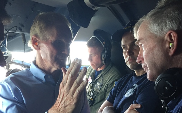 A group of individuals are gathered inside what appears to be a military or Coast Guard aircraft cockpit/cabin area. A civilian in a blue button-down shirt is gesturing with his hand while speaking to several uniformed personnel. The personnel visible include individuals wearing Coast Guard uniforms and aviation headsets.