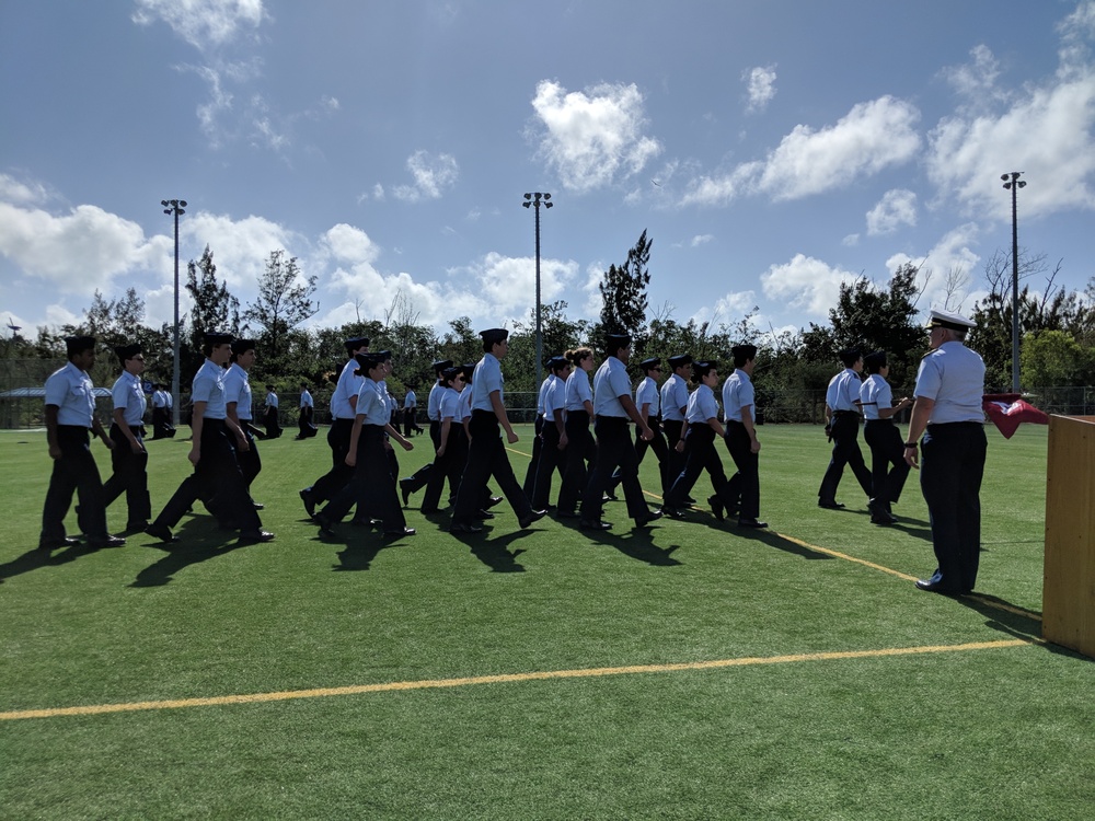 A group of uniformed cadets or JROTC students are marching in formation across an artificial turf athletic field. They are wearing matching white short-sleeve shirts, black trousers, and dark caps. An instructor or senior officer in a white-topped cap stands to the right, observing the formation near what appears to be a podium or reviewing stand.