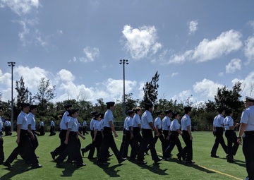 A group of uniformed cadets or JROTC students are marching in formation across an artificial turf athletic field. They are wearing matching white short-sleeve shirts, black trousers, and dark caps. An instructor or senior officer in a white-topped cap stands to the right, observing the formation near what appears to be a podium or reviewing stand.