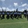 A group of uniformed cadets or JROTC students are marching in formation across an artificial turf athletic field. They are wearing matching white short-sleeve shirts, black trousers, and dark caps. An instructor or senior officer in a white-topped cap stands to the right, observing the formation near what appears to be a podium or reviewing stand.