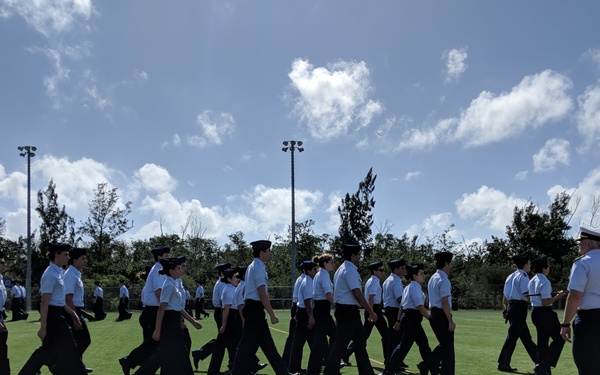 A group of uniformed cadets or JROTC students are marching in formation across an artificial turf athletic field. They are wearing matching white short-sleeve shirts, black trousers, and dark caps. An instructor or senior officer in a white-topped cap stands to the right, observing the formation near what appears to be a podium or reviewing stand.
