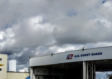 A panoramic view of a U.S. Coast Guard air station facility featuring a large aircraft hangar and an adjacent multi-story administrative building on a tarmac. The hangar doors are open, revealing aircraft and equipment inside. A white air traffic control tower is visible on the right side of the image.
