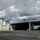 A panoramic view of a U.S. Coast Guard air station facility featuring a large aircraft hangar and an adjacent multi-story administrative building on a tarmac. The hangar doors are open, revealing aircraft and equipment inside. A white air traffic control tower is visible on the right side of the image.