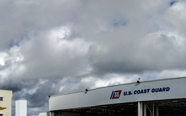 A panoramic view of a U.S. Coast Guard air station facility featuring a large aircraft hangar and an adjacent multi-story administrative building on a tarmac. The hangar doors are open, revealing aircraft and equipment inside. A white air traffic control tower is visible on the right side of the image.