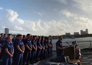 A panoramic photograph taken on the deck of a U.S. Coast Guard vessel, showing a formal ceremony or press event. A large group of uniformed Coast Guard personnel stand in formation while an officer speaks at a podium. Media personnel with cameras are present on the right side, and bales of what appear to be seized contraband are visible in the foreground.