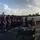 A panoramic photograph taken on the deck of a U.S. Coast Guard vessel, showing a formal ceremony or press event. A large group of uniformed Coast Guard personnel stand in formation while an officer speaks at a podium. Media personnel with cameras are present on the right side, and bales of what appear to be seized contraband are visible in the foreground.