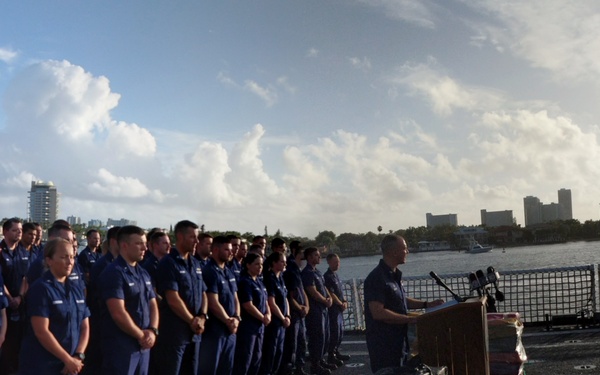 A panoramic photograph taken on the deck of a U.S. Coast Guard vessel, showing a formal ceremony or press event. A large group of uniformed Coast Guard personnel stand in formation while an officer speaks at a podium. Media personnel with cameras are present on the right side, and bales of what appear to be seized contraband are visible in the foreground.