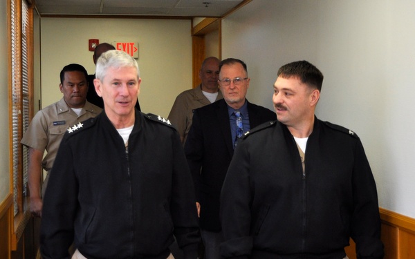 A group of individuals walking down an indoor hallway, with two men in the foreground wearing matching black zip-up jackets and khaki/tan trousers that appear to be military-style uniforms. The man on the left has visible rank insignia (stars) on his shoulders, suggesting senior military officer status. Behind them follow several other individuals, including a person in a tan military uniform and a civilian in a dark suit.