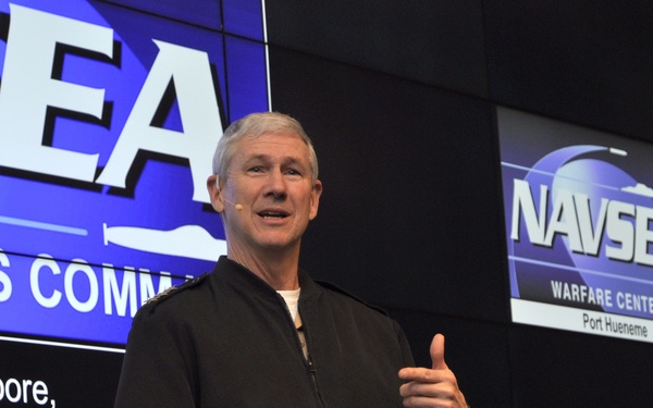 A man is speaking at a NAVSEA (Naval Sea Systems Command) event at Port Hueneme Warfare Centers. He is standing in front of a large display screen showing the NAVSEA logo and partial text identifying 'Thomas J. Moore' of 'Sea Systems Command.' He is wearing a dark zip-up jacket with shoulder insignia and khaki pants, holding note cards while gesturing.