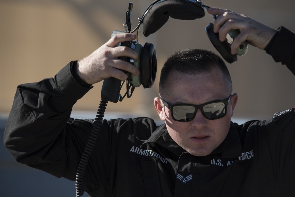 A U.S. Air Force airman named Armstrong is being fitted with or removing a flight/communications headset. He is wearing a black U.S. Air Force uniform with his name tape visible, along with reflective sunglasses. A second person's hands are visible assisting with the headset from above.
