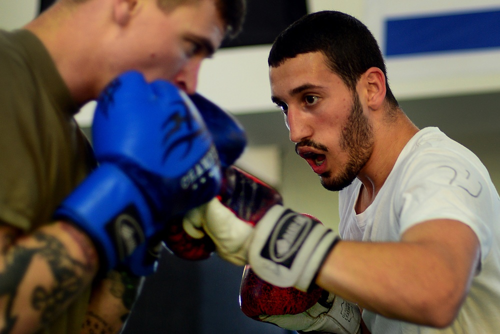 Two individuals are engaged in a boxing sparring session in what appears to be a gym or training facility. The fighter in focus on the right is wearing a white t-shirt and red/white boxing gloves, throwing a punch toward his sparring partner. The partner on the left is wearing blue boxing gloves and an olive-colored shirt, with visible tattoos on his arm.
