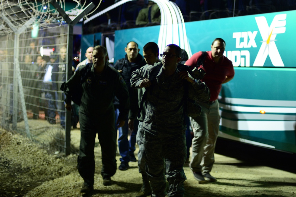A nighttime scene showing a group of individuals, including at least one person in military camouflage uniform, moving along a fenced perimeter near a teal/green bus with Hebrew text. The setting appears to be a secured or controlled area with chain-link fencing topped with razor wire visible on the left. The image was taken in low-light conditions, suggesting a nighttime operation or exercise.