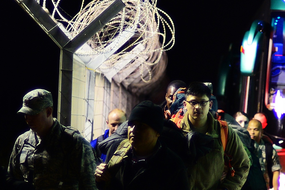 A group of people, including at least one individual in military camouflage uniform, moving at night near a security fence topped with razor/concertina wire. Several individuals are carrying backpacks, and a large vehicle (likely a bus) is visible in the background. The scene appears to take place at a secured facility or checkpoint under artificial lighting.