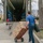 A man in a blue t-shirt and jeans is loading or unloading a large moving truck parked on a residential street. He is using a red hand truck/dolly to transport stacked cardboard boxes, while also carrying a rolled item under his arm. The truck's interior is visible, showing blue moving blankets and other packed items.
