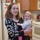 A young woman with brown hair is standing in a residential kitchen holding a young infant dressed in a striped outfit with pink bottoms. Both subjects are facing the camera, with the woman smiling. The kitchen background includes a white refrigerator, white gas stove, microwave, and wooden cabinets.