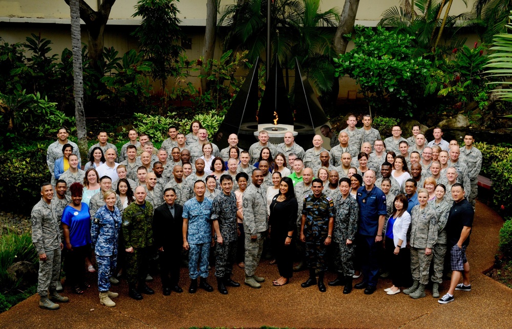 A large group photograph of approximately 100 military personnel and civilians posing together in an outdoor courtyard with tropical vegetation. The group includes members wearing multiple different military uniforms, suggesting a multinational or joint-service gathering. A decorative fountain or flame feature is visible in the background center.