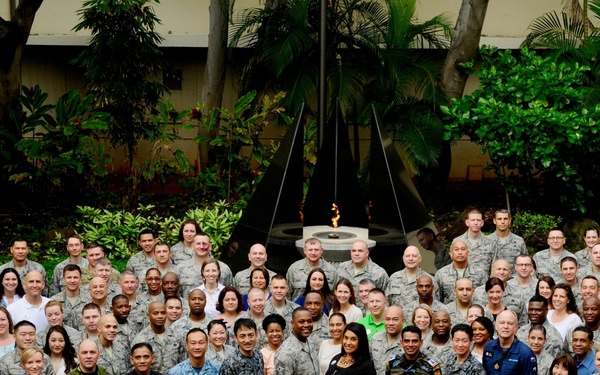 A large group photograph of approximately 100 military personnel and civilians posing together in an outdoor courtyard with tropical vegetation. The group includes members wearing multiple different military uniforms, suggesting a multinational or joint-service gathering. A decorative fountain or flame feature is visible in the background center.