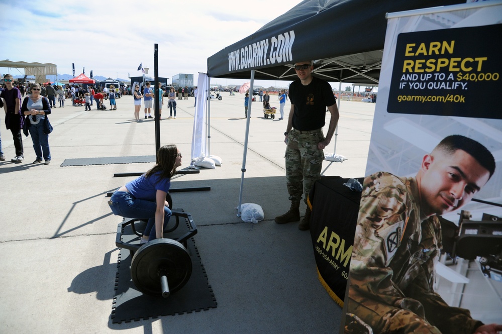 A U.S. Army recruitment booth at what appears to be an outdoor airshow or public event. A young woman in a blue shirt is attempting a deadlift on a barbell station while a soldier in camouflage uniform observes. Recruitment signage prominently displays 'www.goarmy.com' and an incentive banner reading 'Earn Respect and up to $40,000 if you qualify.'