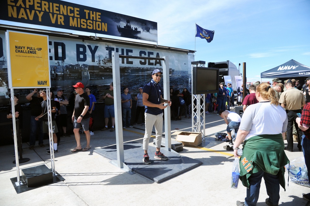 A U.S. Navy recruitment and outreach event is taking place outdoors, featuring interactive stations including a 'Navy Pull-Up Challenge' and a 'Navy VR Mission' experience. A crowd of civilians and military personnel are gathered around the exhibit booths and activity stations. The event appears to be part of a public airshow or military expo, indicated by the professional signage, Navy branding, and large attendance.
