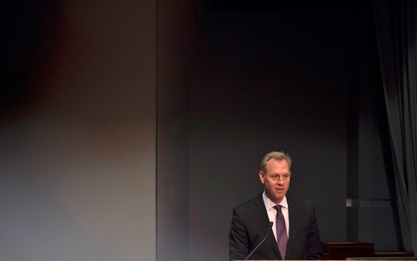 A man in a dark suit and purple tie is speaking at a podium bearing the official seal of the Inspector General, Department of Defense. The setting appears to be a formal auditorium or conference room with dark curtains in the background. The foreground is partially obscured by an out-of-focus element, suggesting the photo was taken from behind an audience member.