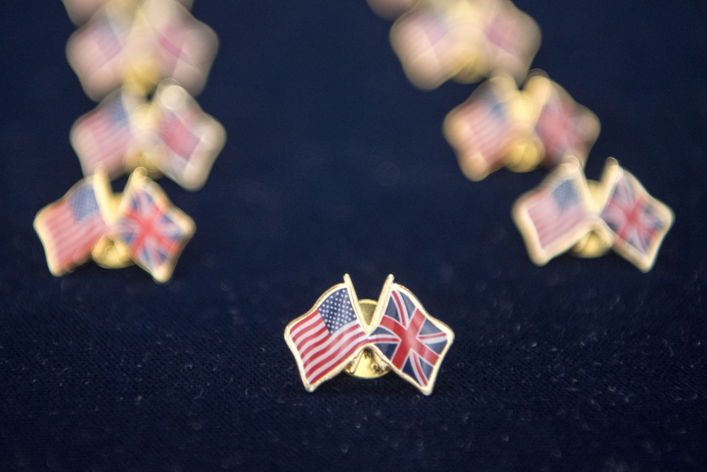 The image shows a collection of dual-flag lapel pins featuring the American (Stars and Stripes) and British (Union Jack) flags crossed together on gold-toned metal bases. One pin is in sharp focus in the center foreground, while multiple identical pins are arranged in a semicircle in the blurred background. The pins are displayed on a dark navy blue fabric surface.