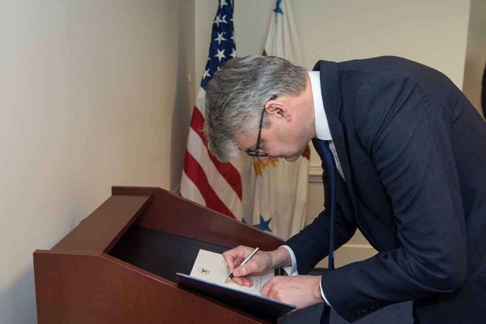A man in a dark suit is leaning over a wooden podium or lectern, signing a document or card with a pen. An American flag and what appears to be a government or military flag are visible in the background. The setting suggests an official or formal ceremony.