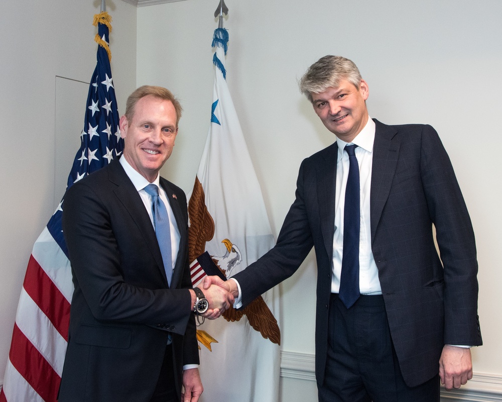 Two men in dark business suits are shaking hands in what appears to be an official government setting. Behind them are two flags: a United States flag and what appears to be a Department of Defense or similar official U.S. government flag featuring an eagle emblem. The setting and flags suggest a formal diplomatic or governmental meeting.