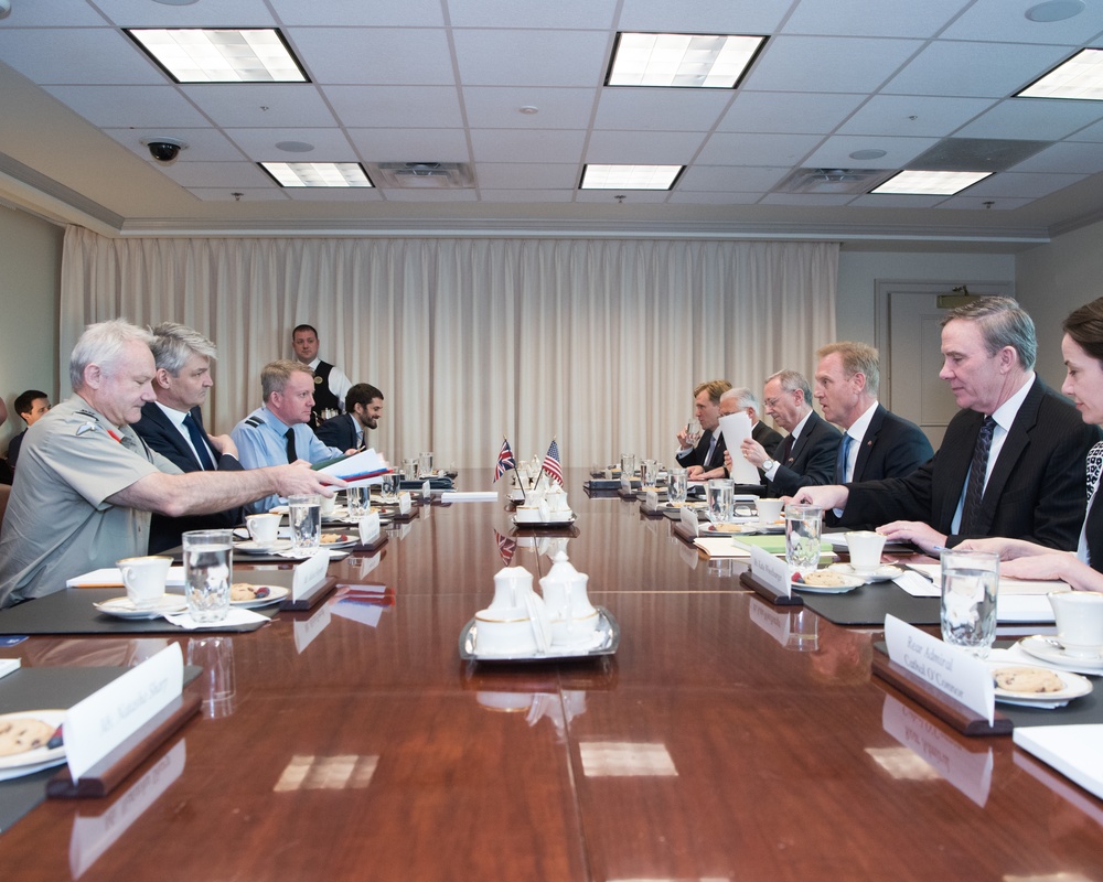 A formal bilateral meeting is taking place around a large polished conference table, with two delegations seated on opposite sides. Small national flags, including a Union Jack and an American flag, are visible at the center of the table, suggesting a US-UK official meeting. Name placards, documents, refreshments, and water glasses are arranged along the table.