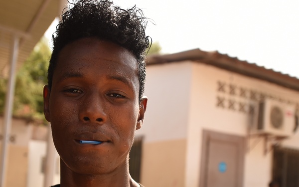 A young man stands outdoors in front of white-painted buildings, holding a blue toothbrush in his mouth. He is wearing a striped t-shirt in gray, yellow, and black. The setting appears to be a compound or institutional facility with multiple structures visible in the background.