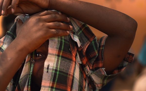 A child is covering their face with their own shirt, pulling it upward to obscure most of their face while peering through with one visible eye. The child wears a worn plaid/checkered shirt in orange, green, and black tones. The background is blurred, showing other figures in an outdoor setting with warm ambient lighting.