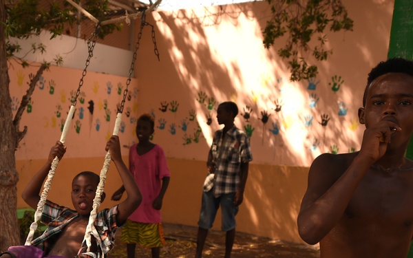 A group of children are playing in an outdoor courtyard area near a tree with a rope-and-chain swing. The background wall is painted in a peach/orange color and decorated with colorful handprint artwork. The scene appears to be at a community center, school, or care facility.