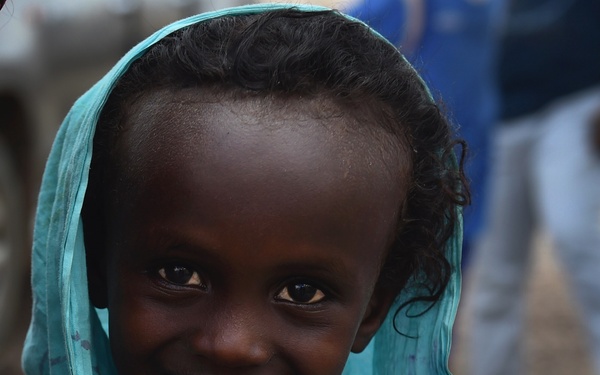 A young child is photographed in a close-up portrait, smiling warmly at the camera while holding a teal/turquoise fabric head covering up to their face. The child appears joyful and candid, wearing layered clothing including a brown garment underneath the teal wrap. The background is blurred, suggesting an outdoor setting with other people or objects present.