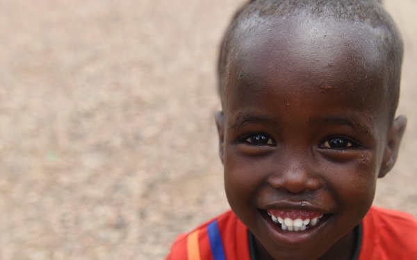 A young child is photographed from a slightly elevated angle, smiling broadly and looking up at the camera. The child is wearing a red t-shirt with yellow and blue stripe detailing. The background is a blurred sandy or gravelly ground surface.