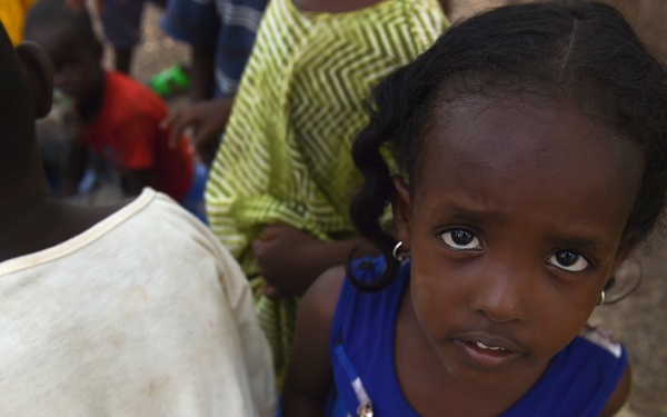 A young girl wearing a blue sleeveless top looks directly at the camera with a serious expression. She is surrounded by other children in what appears to be an outdoor communal setting. The image has a documentary or photojournalistic quality.