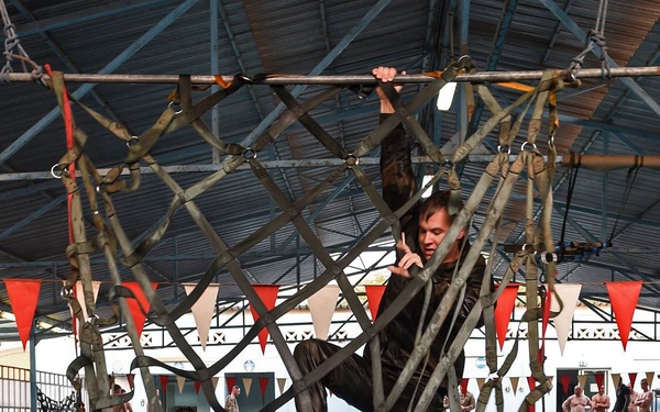 Military personnel are conducting water survival or combat swimming training in an indoor pool facility. One individual is climbing through a large cargo/rope net suspended above the water, while two others are submerged in the pool holding the net from below. Additional personnel and observers are visible in the background near the pool deck.