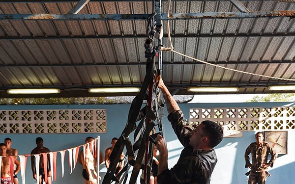 Military personnel in camouflage uniforms are conducting water survival or combat swimming training at an indoor pool facility. Several trainees in the water are climbing a rope net or cargo net suspended from the ceiling, while observers in swimwear and uniformed instructors watch from the pool deck. The exercise appears to simulate helicopter or vessel extraction from water using a net/ladder system.
