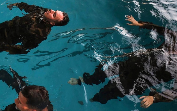 Three individuals wearing camouflage military uniforms are floating or treading water in a pool with clear blue water. The scene appears to depict a military water survival training exercise, as all participants are fully clothed in combat attire. The image is taken from an overhead angle, capturing the rippling water surface and the subjects' positions.