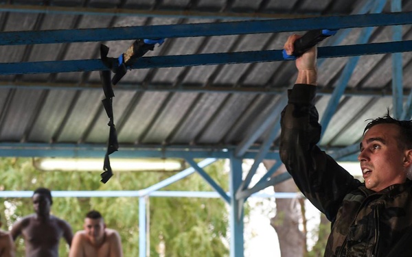 A young man in wet camouflage clothing is performing a hanging bar traverse exercise on blue overhead bars under a covered outdoor structure. Several spectators in swimwear observe from behind a railing in the background. The setting appears to be a military or fitness training facility, possibly near a swimming pool.