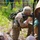 A soldier wearing multicam camouflage uniform, helmet, and a rucksack bearing the number '17' is in a kneeling firing position behind a log barrier with sandbags. Another person is lying on the ground nearby, appearing to simulate a casualty. This appears to be a military training exercise or competition event, evidenced by the numbered identifier and spectators visible behind a yellow boundary rope.