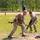 Two soldiers in MultiCam/OCP camouflage uniforms are handling what appears to be a mortar tube during a military competition or training exercise. The soldier in the foreground is numbered '23' and is carrying an M240 machine gun while simultaneously handling the mortar tube. The setting appears to be a marked competition lane on a grassy field with yellow boundary tape and spectators in the background.