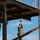 A soldier in camouflage uniform and tactical gear is suspended in mid-air, reaching upward with both hands toward a rope or cable attached to the underside of an elevated wooden and steel platform. The individual appears to be navigating a military obstacle course or rappelling tower, with a rope ladder hanging below their feet. The setting is an outdoor military training facility with pine trees visible in the background under a partly cloudy blue sky.