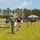 A military competition or training event is taking place on an outdoor grass field, likely a Ranger Challenge or similar Army competition. A soldier in full ACU uniform and helmet wearing the number '1' is running toward the camera, accompanied by a person in a black 'RANGER' t-shirt. Spectators, event tents, portable toilets, and sandbag obstacles are visible in the background.