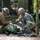 Two U.S. Army soldiers in combat uniforms are providing medical aid to a casualty lying on a litter/stretcher on the ground near a military vehicle. The scene appears to be a training exercise based on the controlled environment, observers in the background, and the staged nature of the scenario. One soldier wears MultiCam (OCP) pattern while the other wears the older ACU (Universal Camouflage Pattern) uniform.