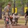 Military personnel are engaged in what appears to be a training exercise in a wooded outdoor environment. One soldier in the foreground wears a number '17' bib and carries a weapon with a red training marker, while another soldier kneels behind a log obstacle in a firing position. A third individual in a black shirt and camouflage pants observes or evaluates the exercise.