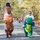 Two participants in a fun run or charity race are wearing inflatable dinosaur costumes — one T-Rex and one green crocodile/alligator — walking along a tree-lined road. A man in a white t-shirt and sunglasses is visible wearing the green inflatable costume from the waist up. Other runners and spectators are visible in the background along the closed road course.