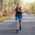 A muscular male runner wearing bib number 199 is competing in an organized road race on a paved road lined with tall pine trees. He is dressed in a black tank top, blue shorts, and blue running shoes, and is wearing sunglasses and earbuds. Other participants are visible in the background further down the road.