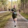 A male runner wearing bib number 1004 and olive green military-style shorts and t-shirt runs along a gravel path flanked by tall pine trees. A second runner is visible in the background, and a road with a vehicle runs parallel to the path. The setting appears to be a organized road race held in a wooded, likely military base environment.